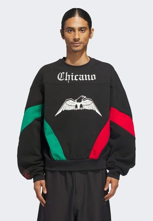 Uomo con capelli lunghi e scuri che indossa una felpa nera con pannelli verdi e rossi, grafica di un'aquila bianca e testo "Chicano" sul petto.