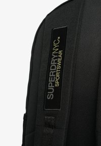 Czarna plecak z teksturowanej tkaniny i wyraźną metką z logo "SUPERDRY NYC SPORTSWEAR" w białych i metaliczno-zielonych literach.