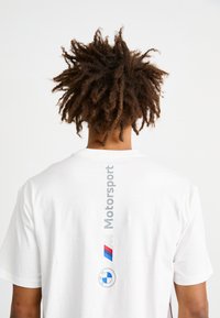 Balta kokvilnas T-kreklā ar pelēko vertikālo uzrakstu "Motorsport", zilo un sarkano BMW logo, un apaļo BMW emblēmu zem tā.