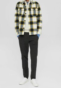 Karierte Flanellhemd in Gelb, Schwarz und Weiß mit zwei Brusttaschen, über einem hellen Pullover getragen. Dunkel karierte Hose und weiße Sneakers.