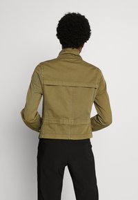 Olivově zelená, přiléhavá bunda s vysokým límcem, přední a zadní yoke a opaskem v pase. Hladká bavlněná tkanina, minimální kovové prvky a přizpůsobené švy.