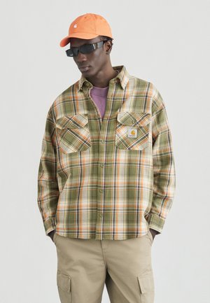 Mees, kes kannab oranži mütsi, musti päikeseprille, rohekas-beezides ruudulise särgiga, mille all lilla särk, ja beežikaid pükse, seisab käed taskutes.