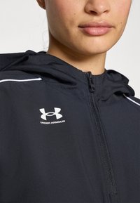 Svart hettejakke laget av glatt materiale, med hel lengde glidelås, hvite detaljer og et Under Armour-logo på fronten.