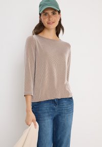 Beige ribbad långärmad topp med rund halsringning, tillsammans med blå jeans. Personen håller en ljusfärgad jacka och bär en grön keps.