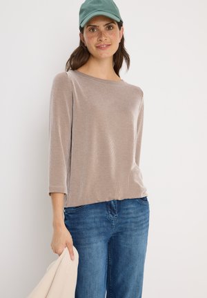 Haut long à manches ribbées beige avec un col rond, associé à un jean bleu. La personne tient une veste claire et porte une casquette verte.