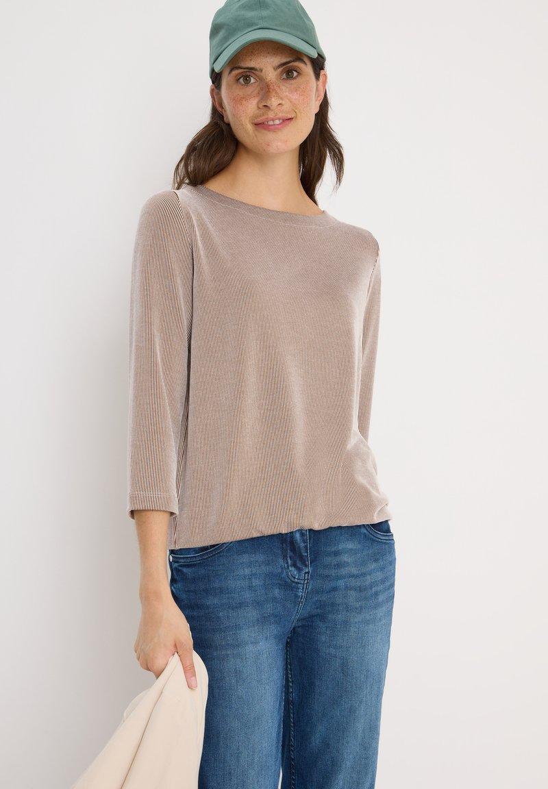 Beige ribbad långärmad topp med rund halsringning, tillsammans med blå jeans. Personen håller en ljusfärgad jacka och bär en grön keps.