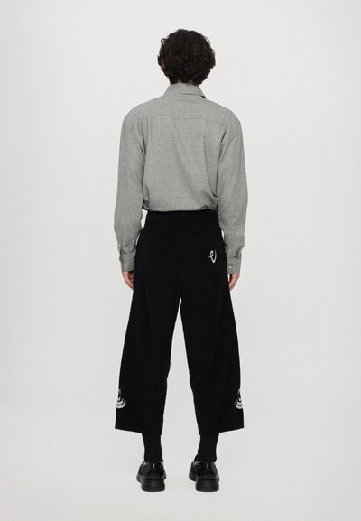 Cămașă gri cu mânecă lungă și guler cu buton, combinată cu pantaloni largi din catifea cu diagonale negre, care au accente brodate pe margini.