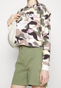 Sudadera corta en un patrón de camuflaje que presenta tonos de verde, rosa y marrón, con un logo blanco. Combinada con pantalones cortos de color verde oliva.