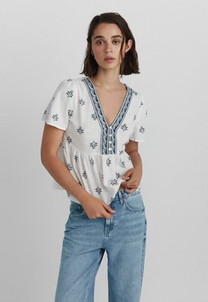 Jonge vrouw draagt een witte blouse met blauwe bloemmotieven en rand, gecombineerd met lichtblauwe spijkerbroek, staand tegen een effen achtergrond.