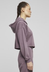 Felpa viola corta realizzata in materiale morbido, con cappuccio regolabile, vestibilità rilassata e polsini a coste. Il modello è mostrato di profilo.