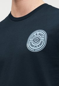 Μπλε ναυτικό βαμβακερό T-shirt με στρογγυλή λαιμόκοψη· διαθέτει ανοιχτό μπλε κυκλικό λογότυπο με κείμενο και λουλουδένιο σχέδιο στο στήθος.