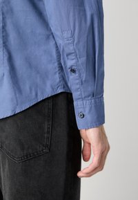 Cămașă butonată de culoare albastră deschisă, cu o textură netedă, având două butoane la manșetă, asortată cu pantaloni negri din denim.