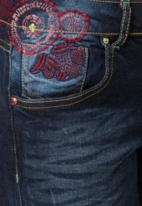 Denim-farkkujen tasku, jossa on kirjailtuja värikkäitä kukkakuoseja punaisella ja violetilla langalla, lähellä keltaisilla ompelemilla saumoilla ja nupiviimeistelyllä.