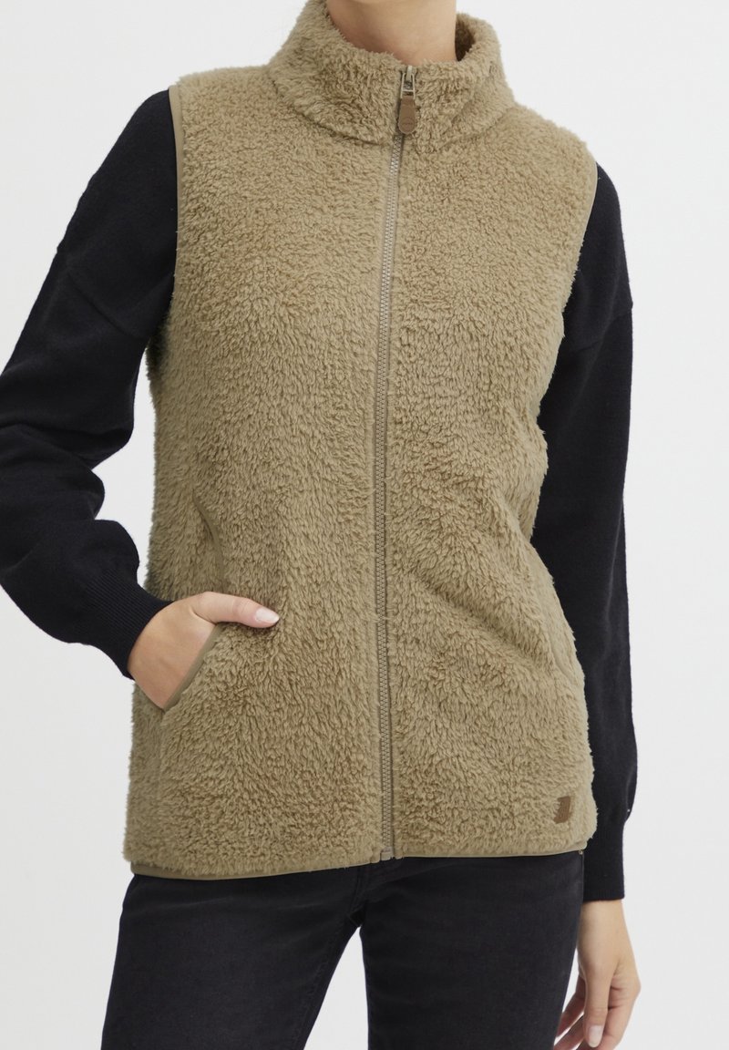 Personne portant un gilet polaire beige zippé sur un t-shirt noir à manches longues, avec une main dans la poche du gilet.