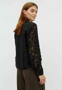 Bluză neagră din material transparent cu model floral, mâneci lungi, manșete butonate și spate complet negru. Puritată peste pantaloni maro.