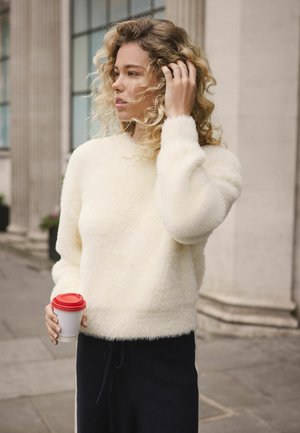 White fluffy sweater with long sleeves, paired with dark navy pants. Person holds a coffee cup with a red lid, standing outdoors.