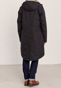 Długi czarny płaszcz z kapturem, z futrzanym kołnierzem, detale z guzikami na rękawach oraz zaokrąglony dół. Nosi się go na ciemnych dżinsach i brązowych butach.