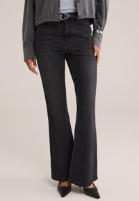 Pantalones vaqueros negros de corte acampanado, hechos de denim, con una cintura alta, diseño clásico de cinco bolsillos y una sutil textura desvaída. Combinados con tacones negros de punta.