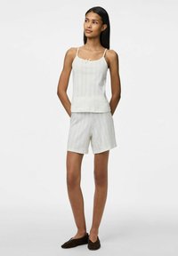 Top blanco sin mangas con tirantes finos, que presenta un pequeño detalle de lazo; combinado con pantalones cortos a juego, ambos en tejido texturizado. Zapatos marrones tipo slip-on.