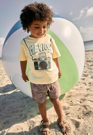 Young child with curly hair standing on sandy beach in sandals, shorts, and yellow t-shirt beside a large multicolored beach ball.