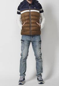Brown puffer vest with a navy top section, white and gray stripe accents, zip closure, paired with distressed blue jeans and sneakers.