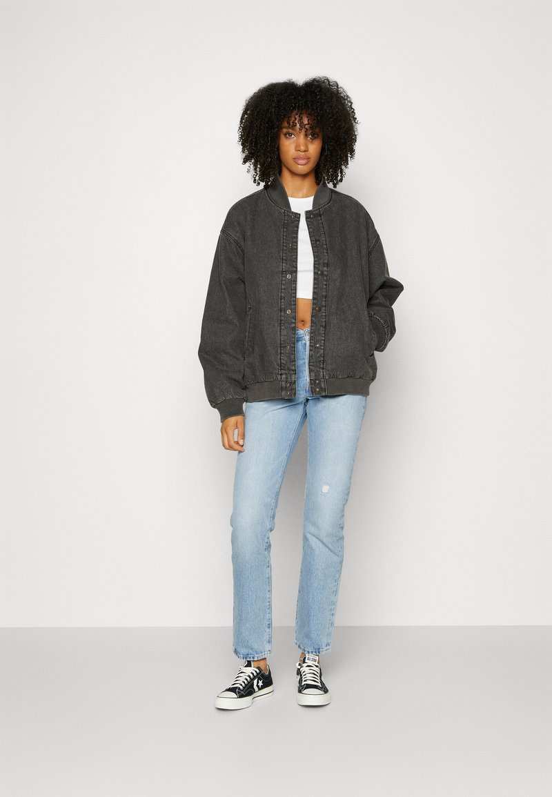 Chaqueta bomber de mezclilla gris oscuro con dobladillo acanalado, llevada sobre un top corto blanco, combinada con jeans rectos de mezclilla azul claro y zapatillas negras.