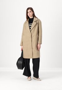 Beige teksturert frakk med notched lapel, sidelommer og en knappelukking, kombinert med svarte vide bukser og beige flats.