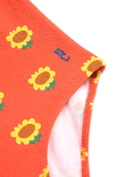 Tissu rouge avec une texture finie, orné de tournesols jaunes et d'un détail bleu en forme de lettre "B" sur le bord. Les bords sont cousus avec soin.