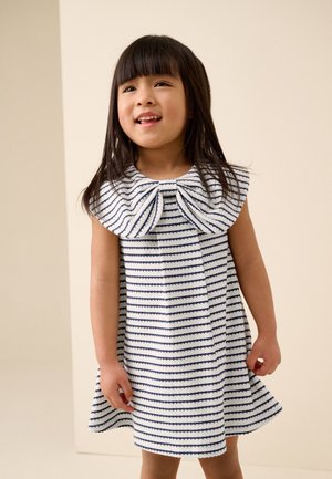 Jong meisje lacht, draagt een witte jurk met marineblauwe horizontale strepen en een grote strik bij de halslijn.