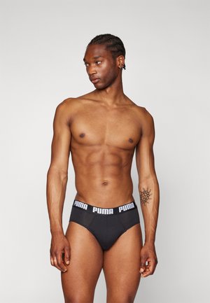 Schwarze Puma Slips mit einem weißen Logo-Band, ausgestattet mit einem figurbetonten Design und glattem Stoff, präsentiert an einem Modell mit durchtrainierter Figur.