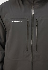 Giacca softshell nera con colletto alto, due tasche sul petto con zip e logo bianco. Progettata per attività all'aperto.