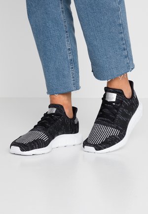 Osoba nosząca czarno-białe prążkowane dzianinowe sneakersy z grubymi białymi podeszwami oraz niebieskie skrócone dżinsy na szarą podłogą.