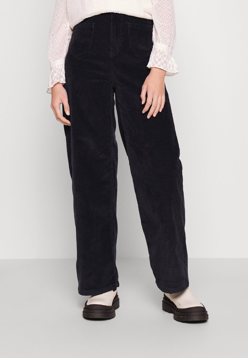 Námořnické kordové kalhoty širokého střihu s dvěma předními kapsami a strukturovaným povrchem, spárované s lehkou blůzou s puntíky a tmavými polobotkami.