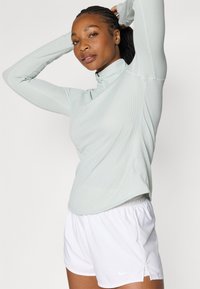 Lys grønne langermede topp med høy krage, teksturert stoff, paret med hvite athletic shorts. Minimalsik design, ingen synlige logoer.