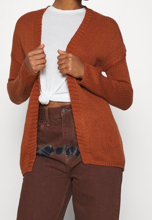 Personne portant un cardigan en maille couleur rouille-orange par-dessus un t-shirt blanc noué et un pantalon marron taille haute avec des taches sombres près de la ceinture.
