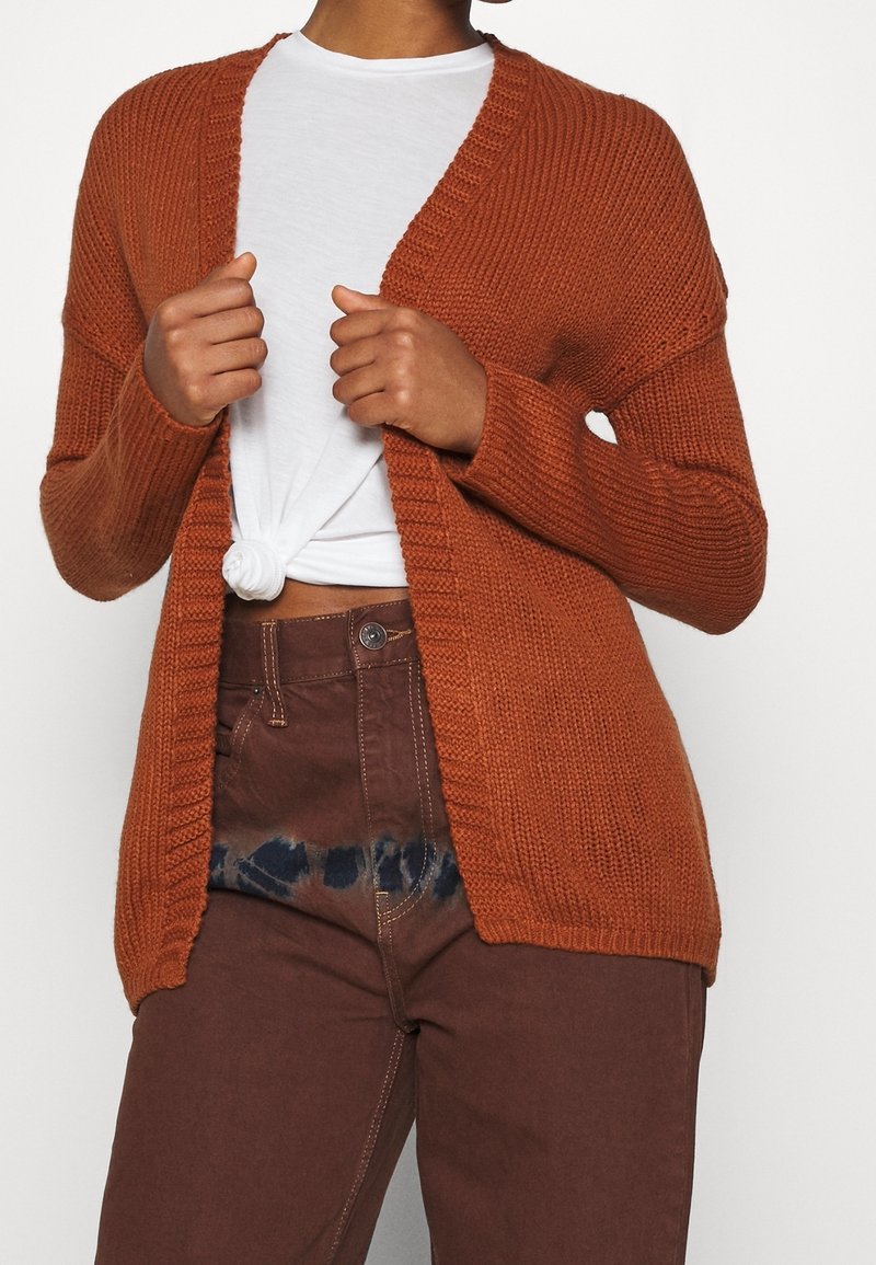 Personne portant un cardigan en maille couleur rouille-orange par-dessus un t-shirt blanc noué et un pantalon marron taille haute avec des taches sombres près de la ceinture.