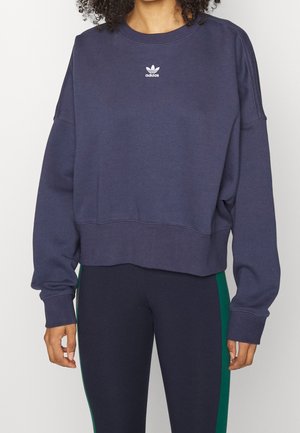 Žena v námořnicky modré mikině Adidas a černých legínách se zelenými pruhy, stojící před jednobarevným bílým pozadím.