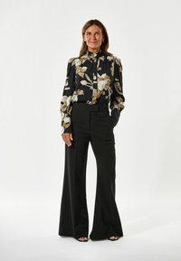Zwarte bloes met bloemeneffect in beige en crème, hoge kraag, lange mouwen. Gecombineerd met zwarte wijde broek met hoge taille.