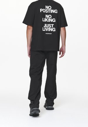 Sort T-shirt med hvid tekst på bagsiden: "NO POSTING NO LIKING JUST LIVING." Kombineret med sorte bukser og sorte sko.