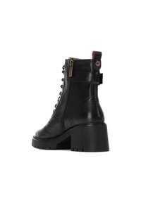Botín de piel negra con tacón grueso, cremallera frontal, detalle de hardware circular y diseño de cordones. Parte superior texturizada con acentos de brogue.