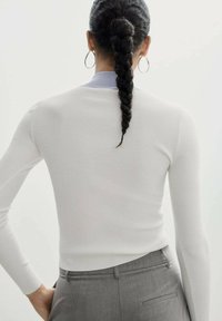 Witte geribbelde langemouwen top met een gebogen zoom, gecombineerd met grijze geklede broek. De top heeft een lichtblauwe kraagaccent.