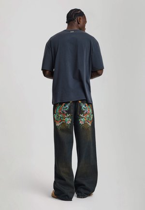 Mann mit geflochtenen Haaren, der ein dunkles, übergroßes T-Shirt und schwarze weit geschnittene Jeans mit bunter Drachenstickerei auf den Gesäßtaschen trägt.