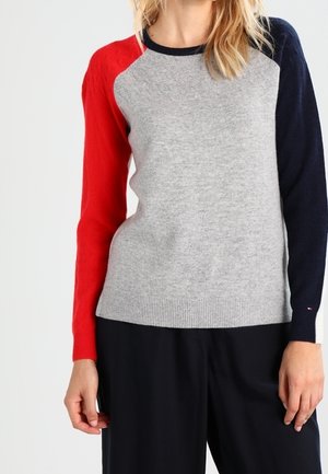 Femme portant un pull gris avec une manche gauche rouge, une manche droite bleu marine, et un pantalon noir sur un fond clair uni.
