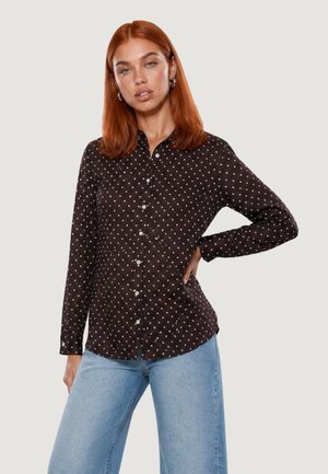 Mujer con cabello rojo que lleva una camisa negra de manga larga con botones y lunares blancos, y vaqueros azul claro, posando con una mano en la cadera.