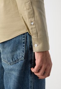 Beige gombos ing mandzsetta gombokkal, kék farmernadrággal párosítva. Az ing sima anyagú és testhezálló, ellentétes textúrákkal.