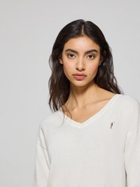 Vit, mjuk tröja med V-ringning och avslappnad passform. Har en liten broderad figur på vänster bröst. Slät textur, minimalistisk design.