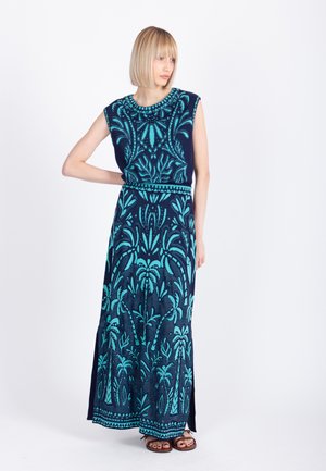 Kvinde iført en ærmeløs maxikjole i marineblåt og teal med palmebladsmotiver, stående med den ene hånd på hoften og kiggende til venstre.