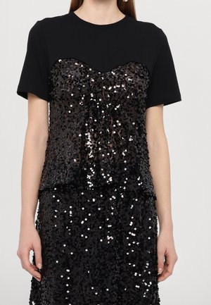 Kvinde iført en sort kortærmet top med en skinnende sort paliet-bustier og matchende nederdel.