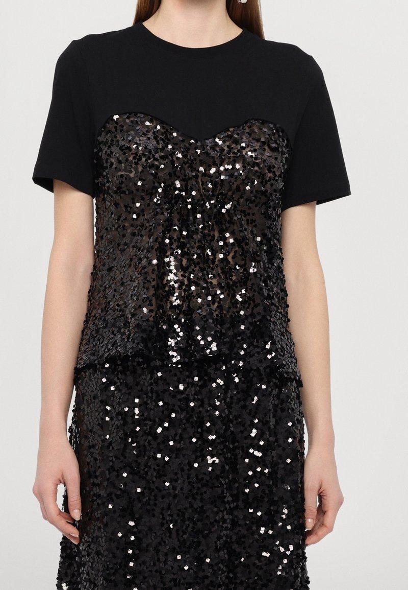 Femme portant un haut noir à manches courtes avec un bustier noir brillant à sequins et une jupe assortie.