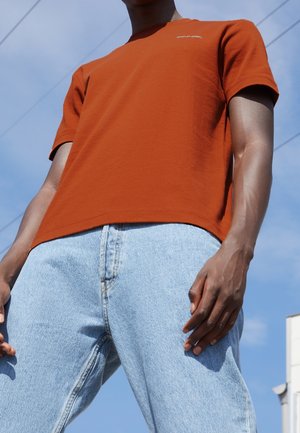 Man met een oranje Calvin Klein T-shirt en lichtblauwe spijkerbroek, staand buiten tegen een achtergrond van een heldere lucht.